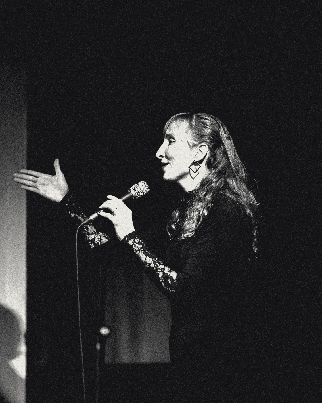 Sylvia in profile on a dark stage, hand outstretched